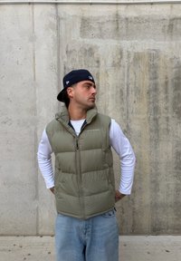 Gilet imbottito verde oliva con collo alto e chiusura a zip, indossato sopra una camicia bianca a maniche lunghe, abbinato a jeans azzurri.