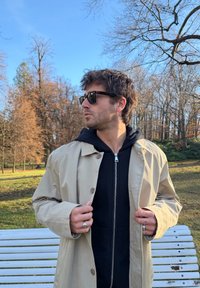 Béžová nepremokavá bunda s veľkými gombíkmi, nosená cez čiernu mikinu na zips. Model má na sebe čierne slnečné okuliare, pozadie tvorí nahé stromy.