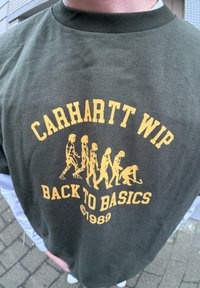 Grøn bomulds T-shirt med gul grafikprint af menneskets evolution. Teksten lyder "CARHARTT WIP BACK TO BASICS ©1989." Rund halsudskæring.