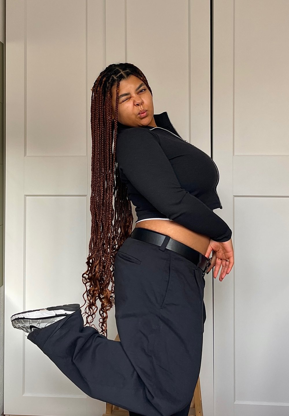 Γυναίκα σε μαύρο κοντό φανελάκι με φερμουάρ και μαύρο παντελόνι, ποζάροντας με το ένα πόδι σηκωμένο. Μακριά, πλεγμένα μαλλιά. Απλό φόντο.