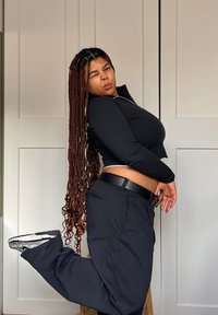 Γυναίκα σε μαύρο κοντό φανελάκι με φερμουάρ και μαύρο παντελόνι, ποζάροντας με το ένα πόδι σηκωμένο. Μακριά, πλεγμένα μαλλιά. Απλό φόντο.