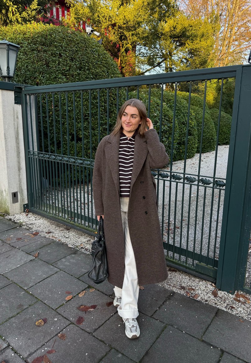 Brown wool coat with a relaxed fit, striped top, light trousers, and grey trainers. Carrying a black handbag, standing by a metal fence.