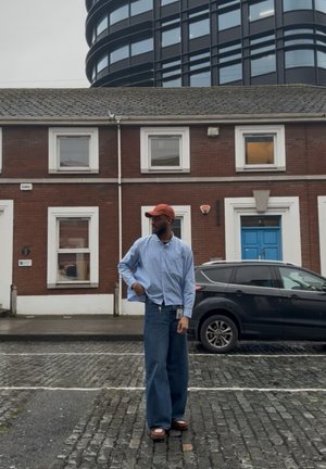 Hombre con gorra naranja, camisa a rayas y vaqueros anchos de pie en una calle empedrada frente a un edificio de ladrillo y un SUV negro estacionado.