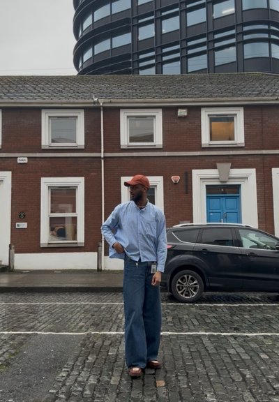 Hombre con gorra naranja, camisa a rayas y vaqueros anchos de pie en una calle empedrada frente a un edificio de ladrillo y un SUV negro estacionado.