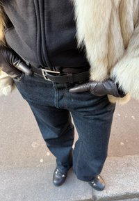 Άτομο που φοράει μαύρα δερμάτινα γάντια, σκούρο μπλε τζιν με μαύρη ζώνη, μαύρο τοπ με φερμουάρ και ανοιχτόχρωμη γούνα.