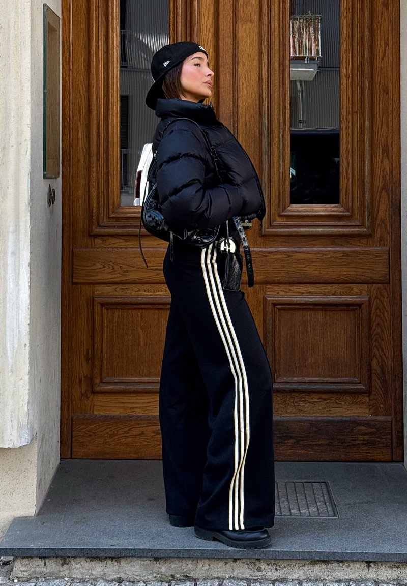 Γυναίκα με μαύρο μπουφάν, μαύρο καπέλο φόρεμα ανάποδα, μαύρο παντελόνι με λευκές ρίγες, μαύρα μποτάκια και μικρό μαύρο σακίδιο στέκεται μπροστά σε ξύλινη πόρτα.