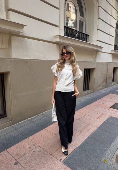 Top blanco texturizado con mangas con volantes, combinado con pantalones anchos negros. Complementado con zapatos planos en blanco y negro y un bolso blanco.