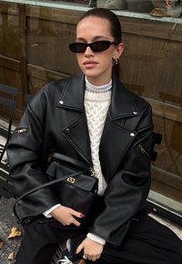 Musta nahkainen ylisuuri takki, jossa vetoketjulliset taskut, puettuna kermanvärisen palmikkoneuleen päälle. Pieni musta laukku kullanvärisillä yksityiskohdilla, tummat aurinkolasit.