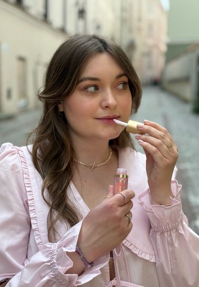 Brillo labial en un tubo transparente con acentos dorados sostenido en una mano, mientras se aplica con un aplicador en la otra; vistiendo una blusa rosa pálido con volantes.