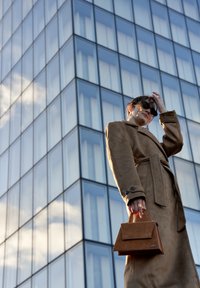 Beige suède trenchcoat met ceintuur in de taille, oversized revers en twee knopen. Houdt een kleine, rechthoekige bruine handtas met klepsluiting vast.