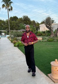 Röd kortärmad skjorta med svart blommönster, svarta byxor och mörka skor. Utomhusmiljö med grönska och en stenlagd väg.