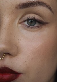 Närbild av ett ansikte med ljusbrun hy, klara gröna ögon, djärva röda läppar och en geometrisk septumring i guld, med välformade ögonbryn.