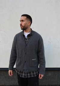 Cardigan gris en maille avec un col en V, une fermeture à boutons et des poches avant. Superposé avec une chemise à carreaux noir et blanc avec un col arrondi.