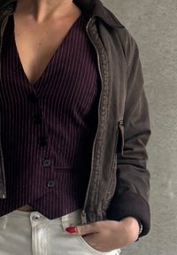 Giacca bomber marrone con zip sopra un gilet a righe bordeaux scuro con bottoni, abbinata a pantaloni bianchi. Il tessuto ha una texture liscia.