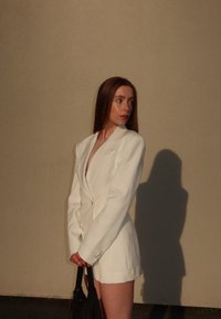 Hvit skreddersydd blazer med brede klaffer og dobbeltraderte knapper, kombinert med svart håndveske, mot en nøytral veggbakgrunn.