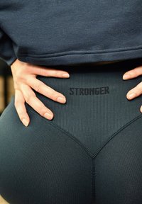 Hender hvilende på hoften over mørke, teksturerte leggings med "STRONGER" trykt på den høye midjen, kombinert med en mørk topp.