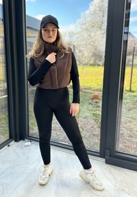 Hnědý prošívaný vestu spárovanou s černým dlouhým rukávem a legínami, doplněnou světlemi teniskami. Pozadí tvoří velká skleněná okna.