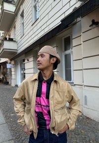 Beige jas van kunstleer met elastische manchetten, gedragen over een zwart poloshirt met een roze kruispatroon en logo. Donkere spijkerbroek zichtbaar.