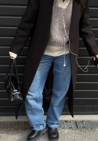 Osoba w długim czarnym płaszczu, w paskowanym swetrze, niebieskich dżinsach i czarnych botkach, trzymająca czarną torebkę i smartfon z słuchawkami.