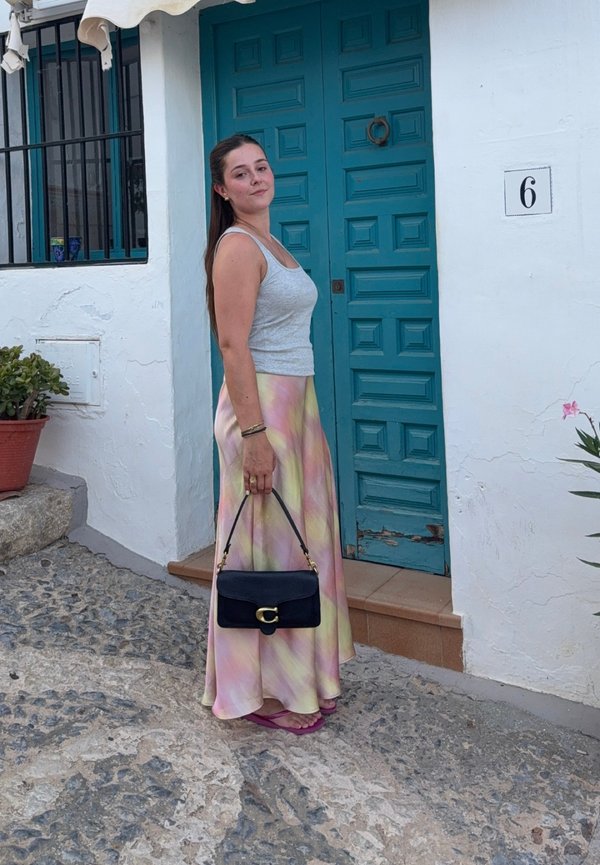 Frau in einem grauen Tanktop und pastellfarbenem Rock steht auf einer Kopfsteinpflasterstraße und hält eine schwarze Handtasche neben einer blauen Tür mit der Nummer sechs.