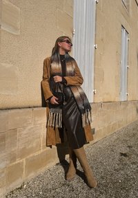 Tan-naha nahast mantel, must sall gradient-mustriga, pruun top ja nahast seelik. Beežid põlveni kõrged saapad, must käekott. Seisab vastu seina.