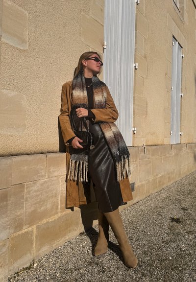Abrigo de ante color tostado, bufanda negra con un patrón degradado, blusa marrón y falda de cuero. Botas beige hasta la rodilla, bolso negro. De pie contra una pared.