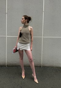 Beige neuletoppi, valkoinen miniönkkä, jossa on sivureikä, ja nude stiletto-korkokengät. Pitelee aikakauslehteä; taustalla harmaa teksturoitu seinä.