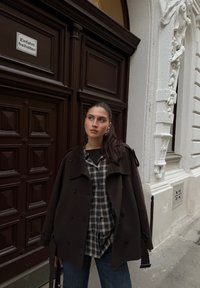 Manteau brun foncé surdimensionné porté sur une chemise à carreaux ; le mannequin se tient à côté d'une porte brune avec une architecture décorative blanche en arrière-plan.