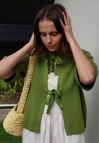 Vihreä neulepusero, jossa on lyhyet hihat ja sitomisella suljettava etuosa. Yhdistetty valkoiseen mekkoon ja korostettu punottu beige laukku.