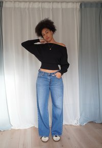 Maglione nero corto con design a scollo off-shoulder, abbinato a jeans a gamba larga color azzurro chiaro. Scarpe da ginnastica casual completano l'outfit.