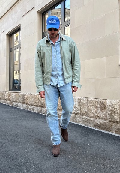 Hombre con gorra azul, gafas de sol, chaqueta verde claro, camisa de mezclilla y jeans, caminando por la acera de una ciudad junto a un edificio de piedra beige.