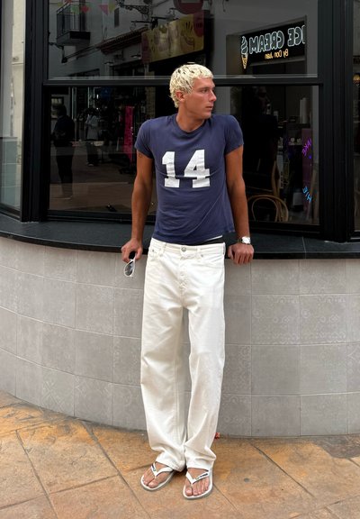Camiseta azul marino con el número 14, pantalones anchos blancos y chanclas. Tiene el cabello corto y rubio, y sostiene unas gafas de sol. Entorno urbano.