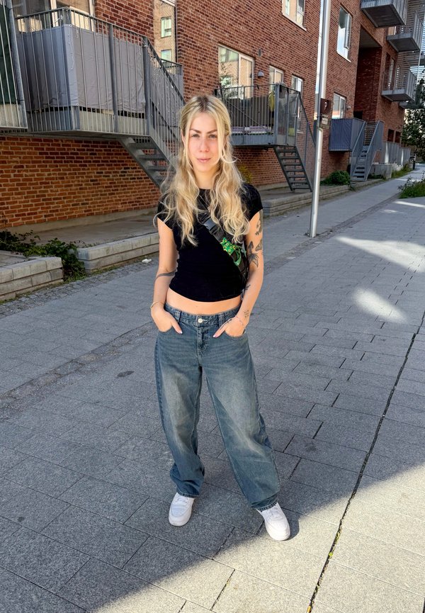 Joven con pelo largo y rubio, lleva un top negro corto, vaqueros holgados y zapatos blancos, está de pie en una calle adoquinada con edificios de ladrillo al fondo.