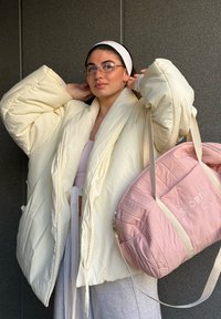Femme ajustant ses lunettes, portant une doudoune crème oversize et un bandeau blanc, portant un sac de voyage matelassé rose avec le texte "Sport Club".