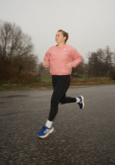 Chaqueta de correr rosa, leggings negros y zapatillas deportivas azules con detalles en blanco. Un desenfoque de movimiento sugiere una carrera activa en una carretera de asfalto gris.