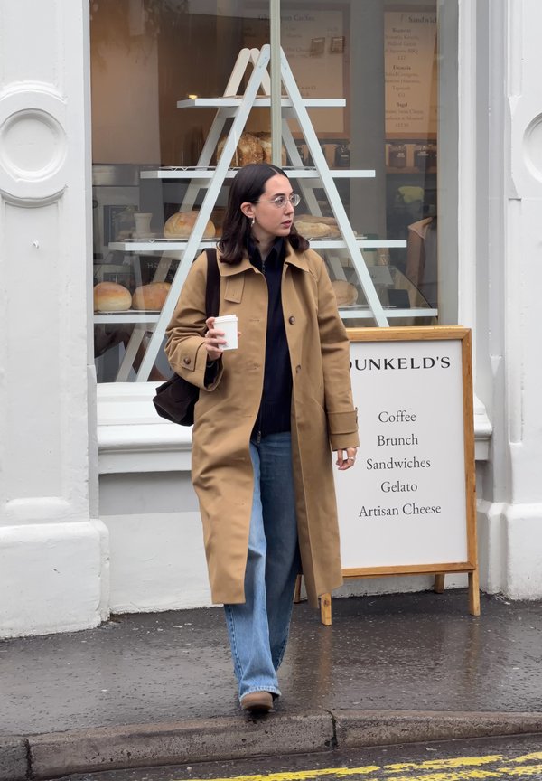 Mujer con abrigo marrón claro y gafas, sosteniendo una taza de café, caminando sobre un pavimento mojado frente a una panadería con pan y un cartel de menú.