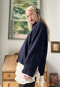 Mulher loira com batom escuro, vestindo um suéter azul-marinho sobre uma saia branca em camadas com volantes, de pé dentro de casa, perto de móveis de madeira e arte.