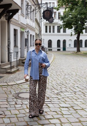Camisa de botones de manga larga de color azul claro con dos bolsillos frontales; pantalones de pierna ancha con estampado de leopardo; sandalias negras; sostiene una taza de café. Fondo de calle de adoquines.