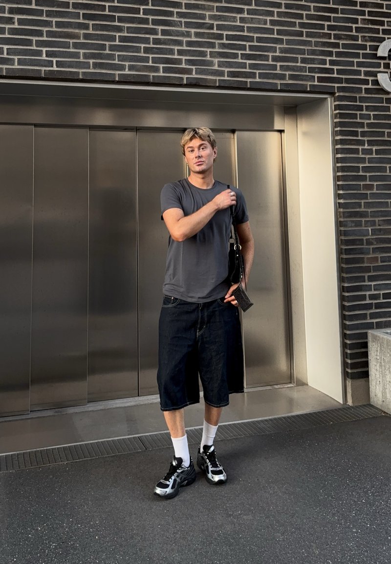 Jeune homme en t-shirt gris et shorts longs se tient à l'extérieur près de portes d'ascenseur en métal, tenant un petit sac sur une épaule.