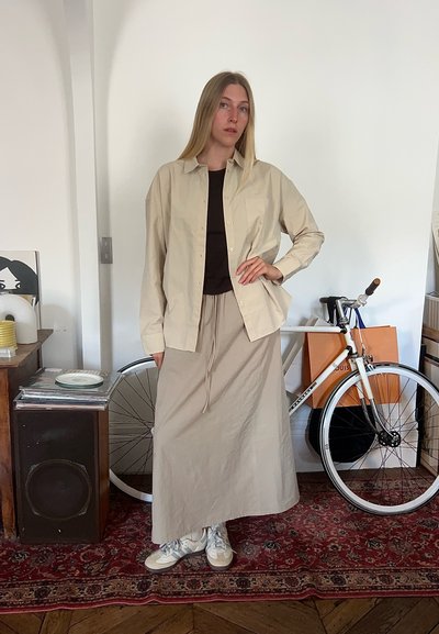 Camisa de botones color beige claro sobre una blusa oscura, combinada con una falda larga a juego. Zapatillas blancas. El fondo incluye una bicicleta y elementos decorativos.