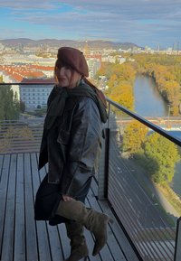 Smeđa kožna jakna, zeleni šal, smeđi beret, crna haljina i čizme do koljena u maslinastoj boji. Pogled s balkona sa gradom i rijekom u pozadini.