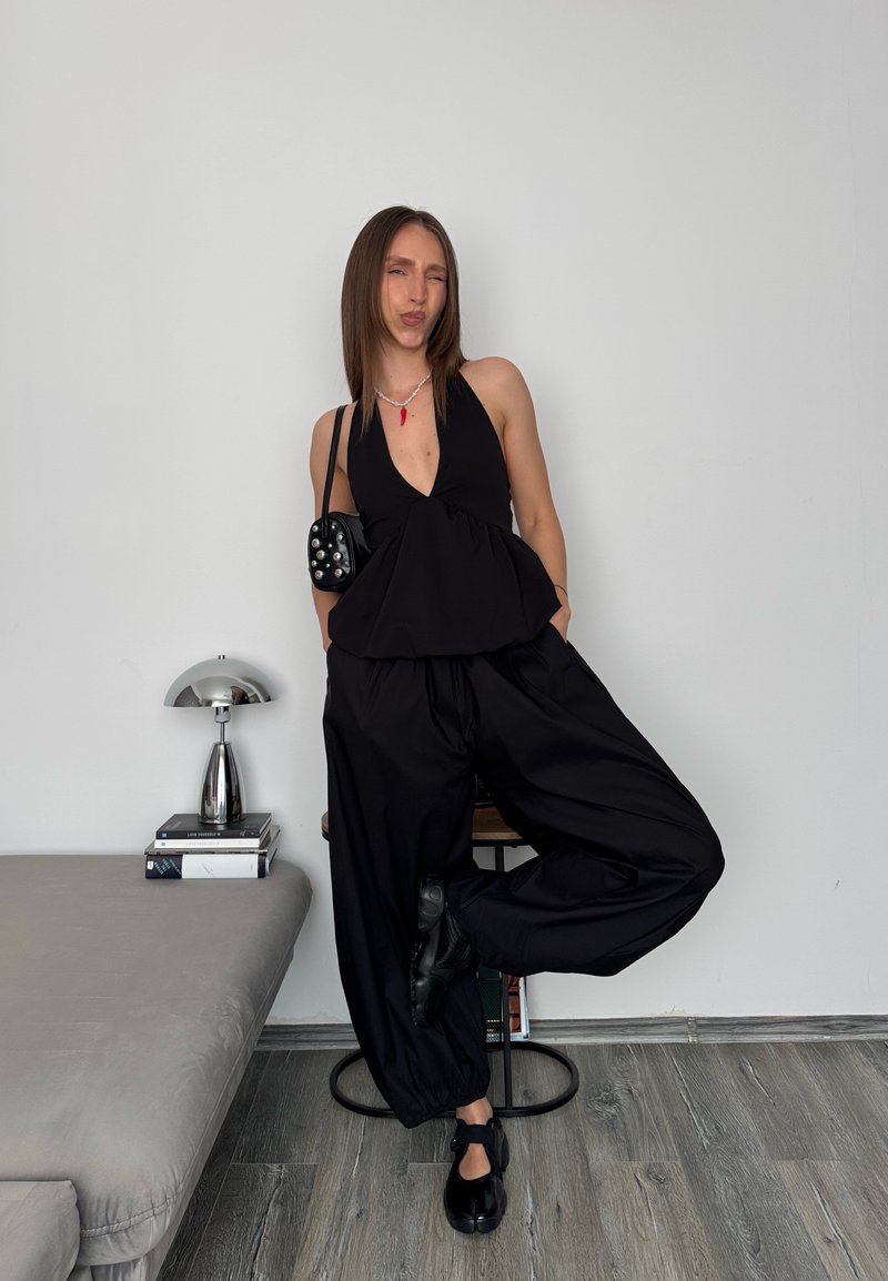 Femme en haut à col halter noir et pantalon posant avec une jambe levée, debout dans une pièce minimaliste avec une lampe et des livres empilés.
