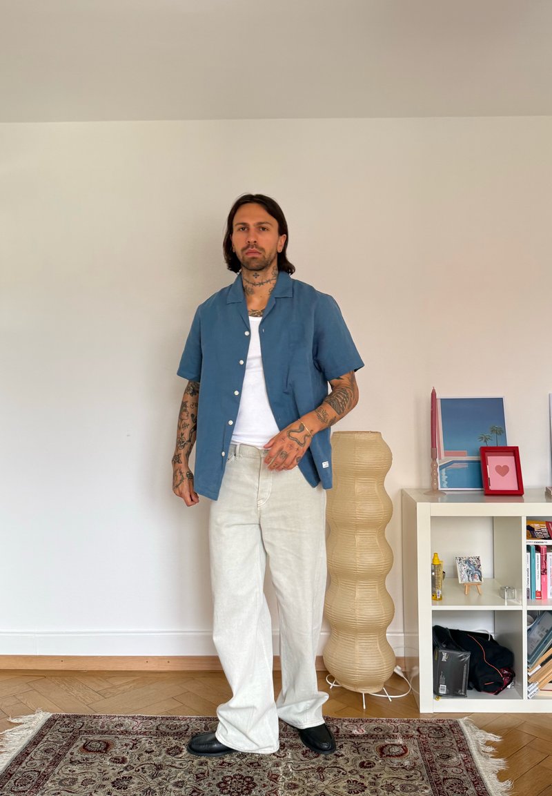 Homme avec des tatouages portant une chemise bleue décontractée, un débardeur blanc et un pantalon blanc ample dans une pièce avec un tapis à motifs, une étagère et une lampe haute.
