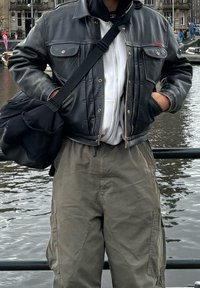 Black leather jacket featuring two chest pockets, a white hooded sweatshirt underneath, and grey cargo trousers. The individual is holding a black bag.