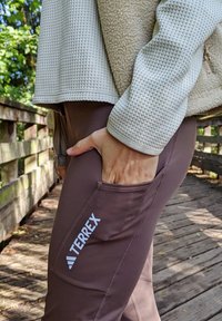 Braune Leggings mit Seitentasche und weißem "TERREX"-Logo; kombiniert mit einem strukturierten, hellgrauen Oberteil mit Fleece-Akzenten.