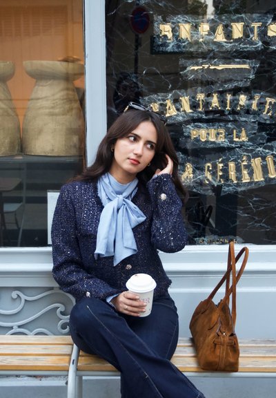 Chaqueta azul marino texturizada con motas doradas, bufanda azul claro, vaqueros oscuros de mezclilla, sosteniendo una taza de café, bolso de ante marrón al lado en el banco.