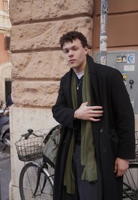 Νέος άνδρας με μαύρο παλτό και πράσινο κασκόλ στηρίζεται σε πέτρινο τοίχο δίπλα σε ποδήλατο με καλάθι σε πολυσύχναστο δρόμο της πόλης.