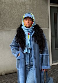 Μακρύ μπλε παλτό με υφή από γούνα και λεπτομέρειες από τεχνόδερμα, φορεμένο πάνω απ ό τζιν. Συμπληρώνεται από μπλέ φουλάρι και καπέλο με φωτεινά χρώματα.