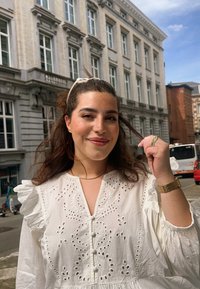 Λευκό κεντημένο φόρεμα με ρανίδες μανίκια, λεπτομέρειες από κουμπιά και σχέδια κοπής; φορεμένο με ένα χρυσό ρολόι και κορδέλα μαλλιών.