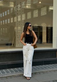 Μια γυναίκα που φοράει μια μαύρη αμάνικη μπλούζα και λευκά παντελόνια με φαρδιά πόδια στέκεται μπροστά από μια βιτρίνα καταστήματος, κρατώντας μια καφέ τσάντα και φοράει γυαλιά ηλίου.